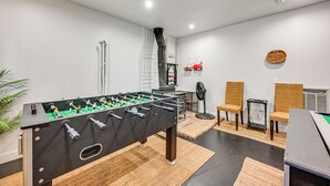 Quarto com mesa de futebol, duas cadeiras de vime, um ventilador e uma pequena mesa com uma placa.