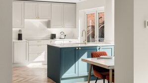 Una cocina moderna con armarios blancos y color turquesa, un comedor con mesa y sillas, y una ventana con vistas a un edificio de ladrillo.