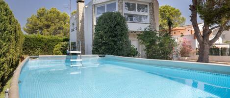 Una piscina con escalera, rodeada de árboles y un edificio de paredes de piedra y ventanas.
