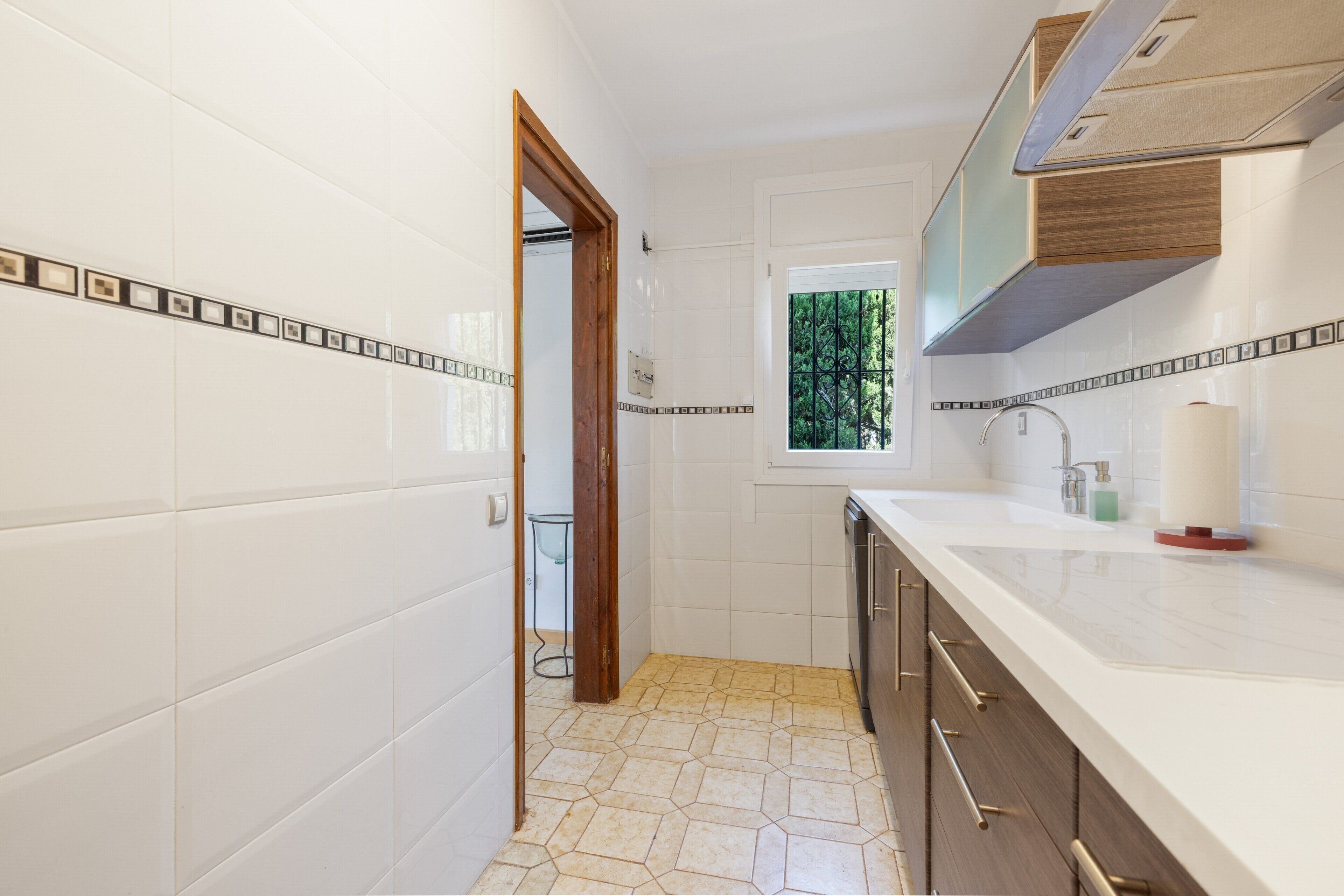 A bathroom with white tiles, a wooden door, a window with a view, and a sink with a mirror.