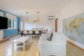 Ein modernes Wohnzimmer mit Sofa, Couchtisch, Esstisch mit Stühlen, Pendelleuchten, einem Flachbildfernseher und einer Weltkarte an der Wand.