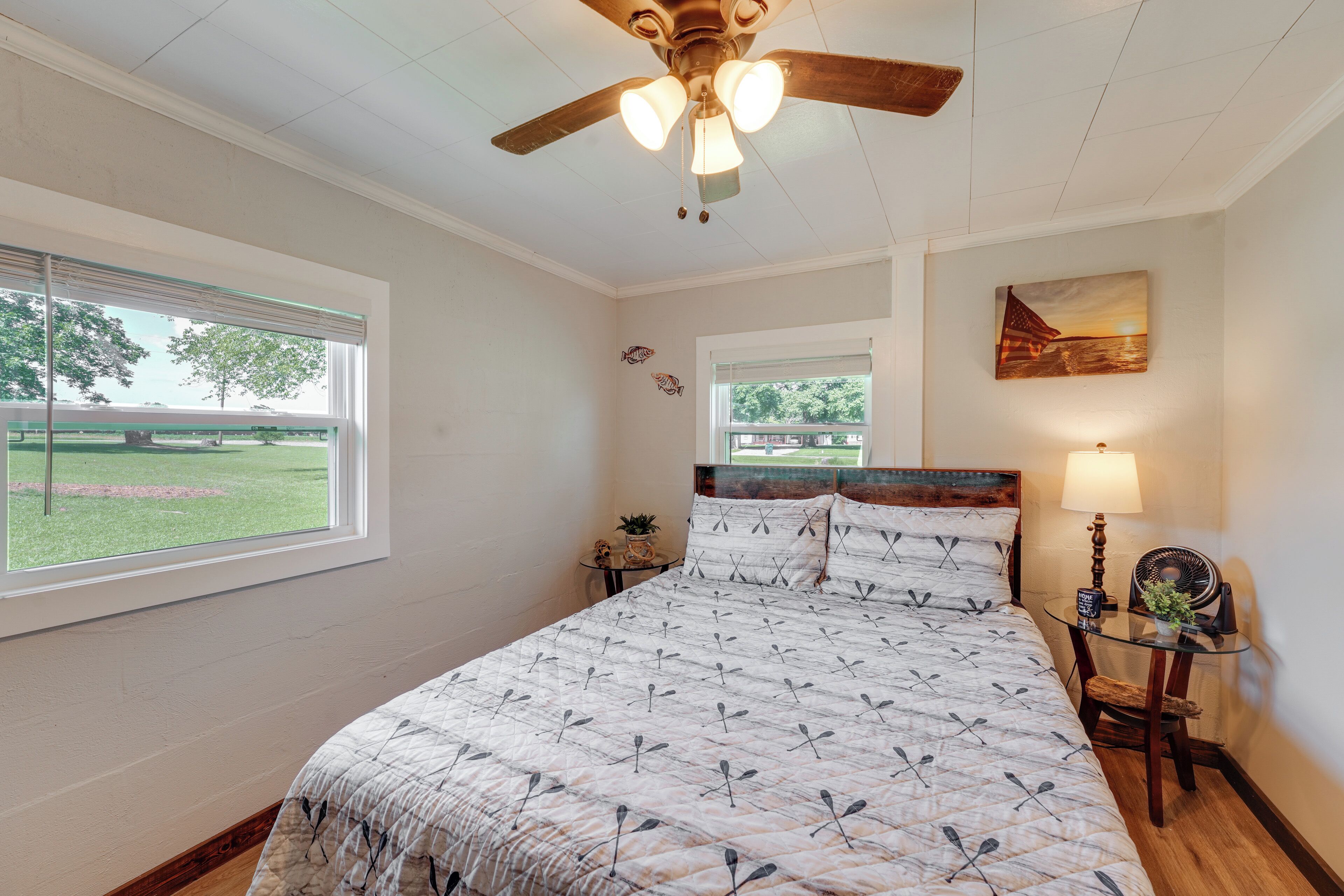 A bedroom with a bed, a ceiling fan, a window, a nightstand, a lamp, and a painting on the wall.