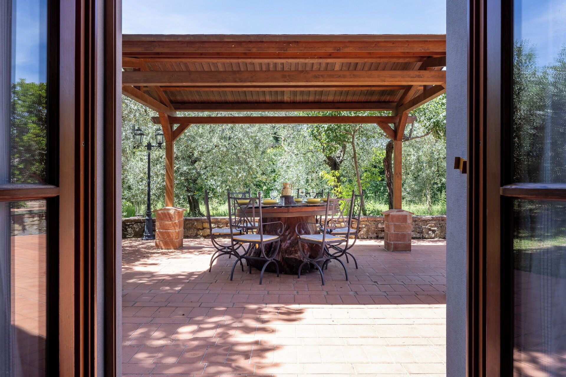 A covered patio with a table and chairs, a stone wall, and trees in the background.