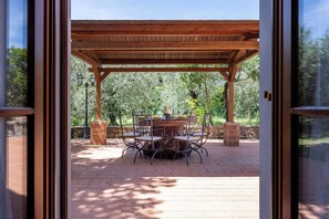 Un patio cubierto con una mesa y sillas, un muro de piedra y árboles al fondo.