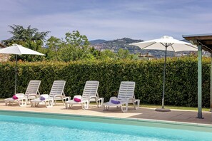 Ein Poolbereich mit Liegestühlen und Sonnenschirmen, einer Hecke und Blick auf eine Stadt im Hintergrund.