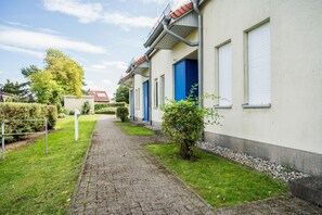 Un chemin mène à une rangée de bâtiments aux portes bleues, entourés de verdure et sous un ciel dégagé.