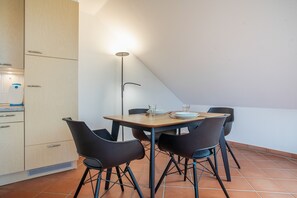 Un coin repas avec une table dressée pour quatre personnes, entourée de chaises, dans une pièce dotée d’un plafond en pente et d’un espace cuisine situé à gauche.