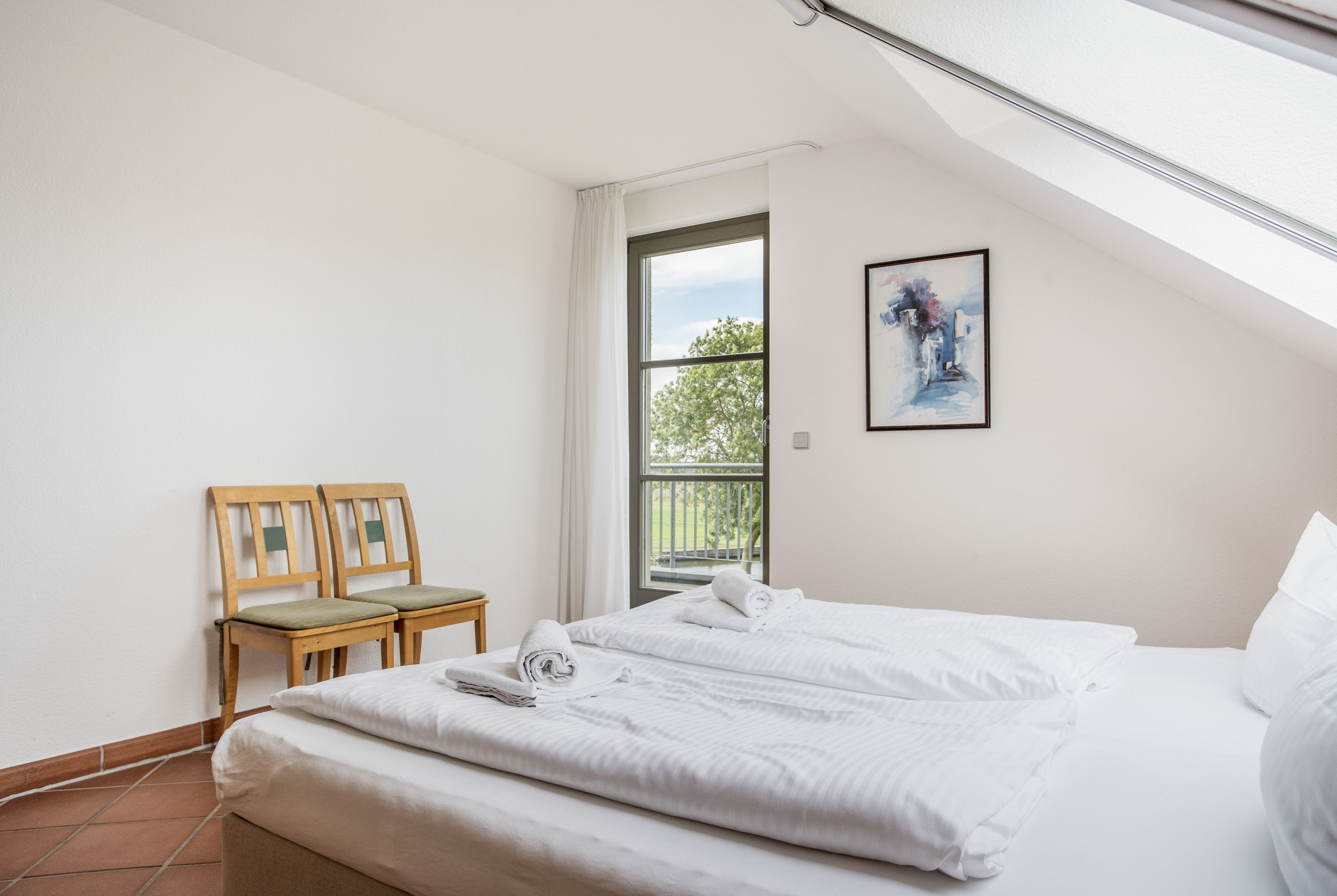 Un dormitorio con cama, ventana, un cuadro y una silla.