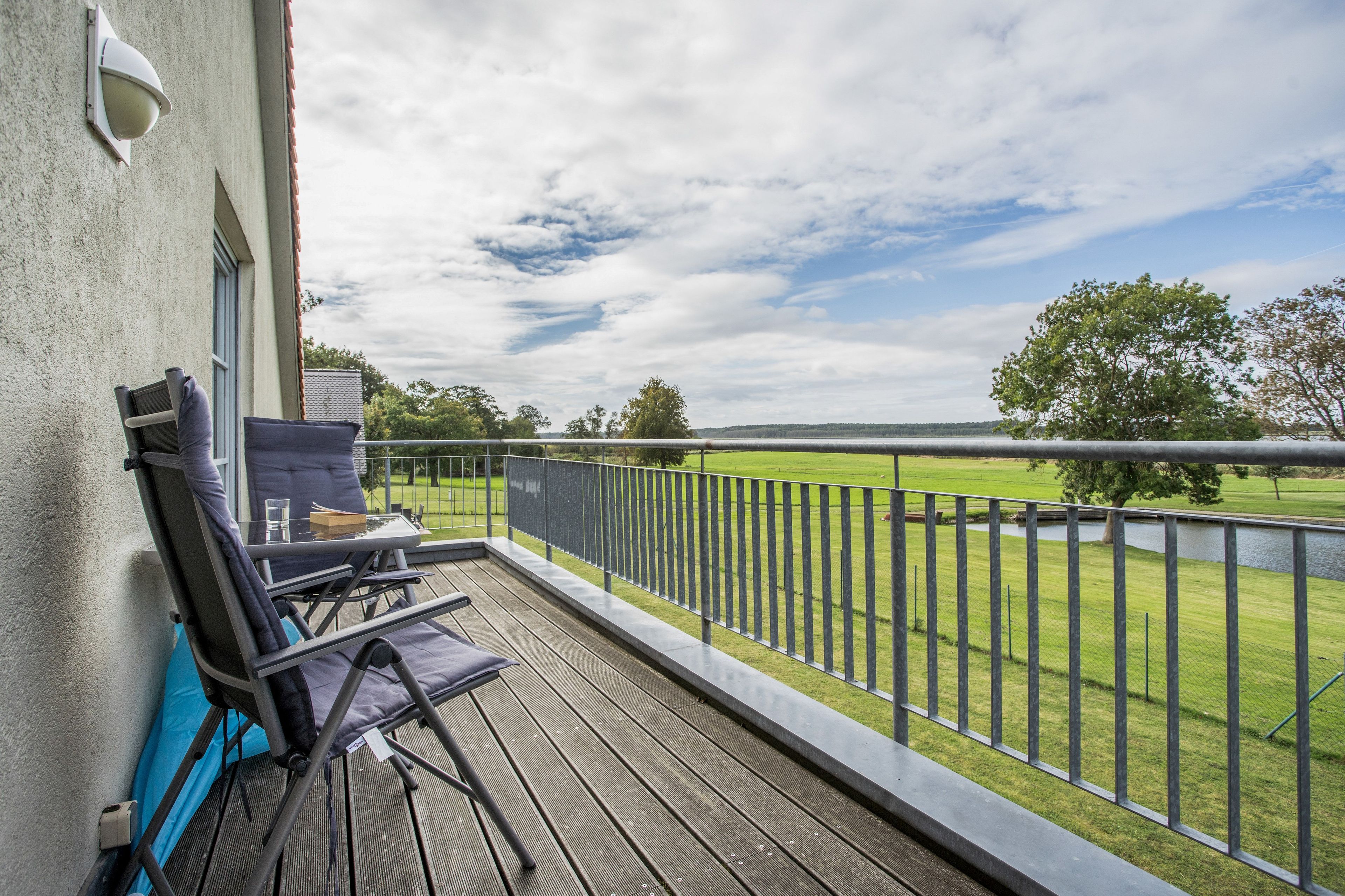 A balcony with a view of a grassy field and trees, a chair, and a table with a glass and a book.