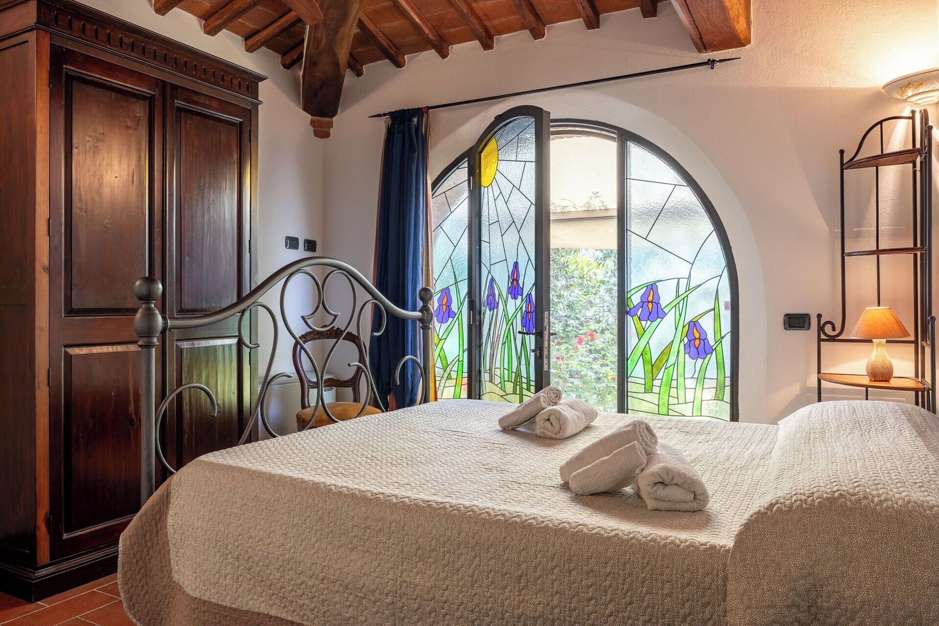 A bedroom with a bed, a wooden wardrobe, a metal bed frame, a window with a stained glass view, and a bedside lamp.