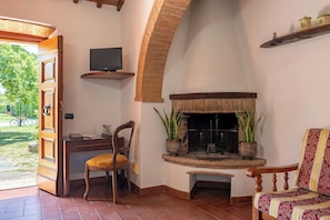 A room with a fireplace, a television on a shelf, a desk with a chair, and a view of the outdoors. - Il Cipresso, Casa Martina, Cortona (Cortona)