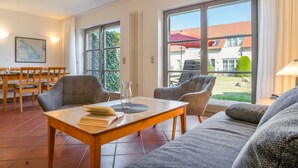 En moderne stue med sofa, stole, spisebord og udsigt til et hus gennem store vinduer.