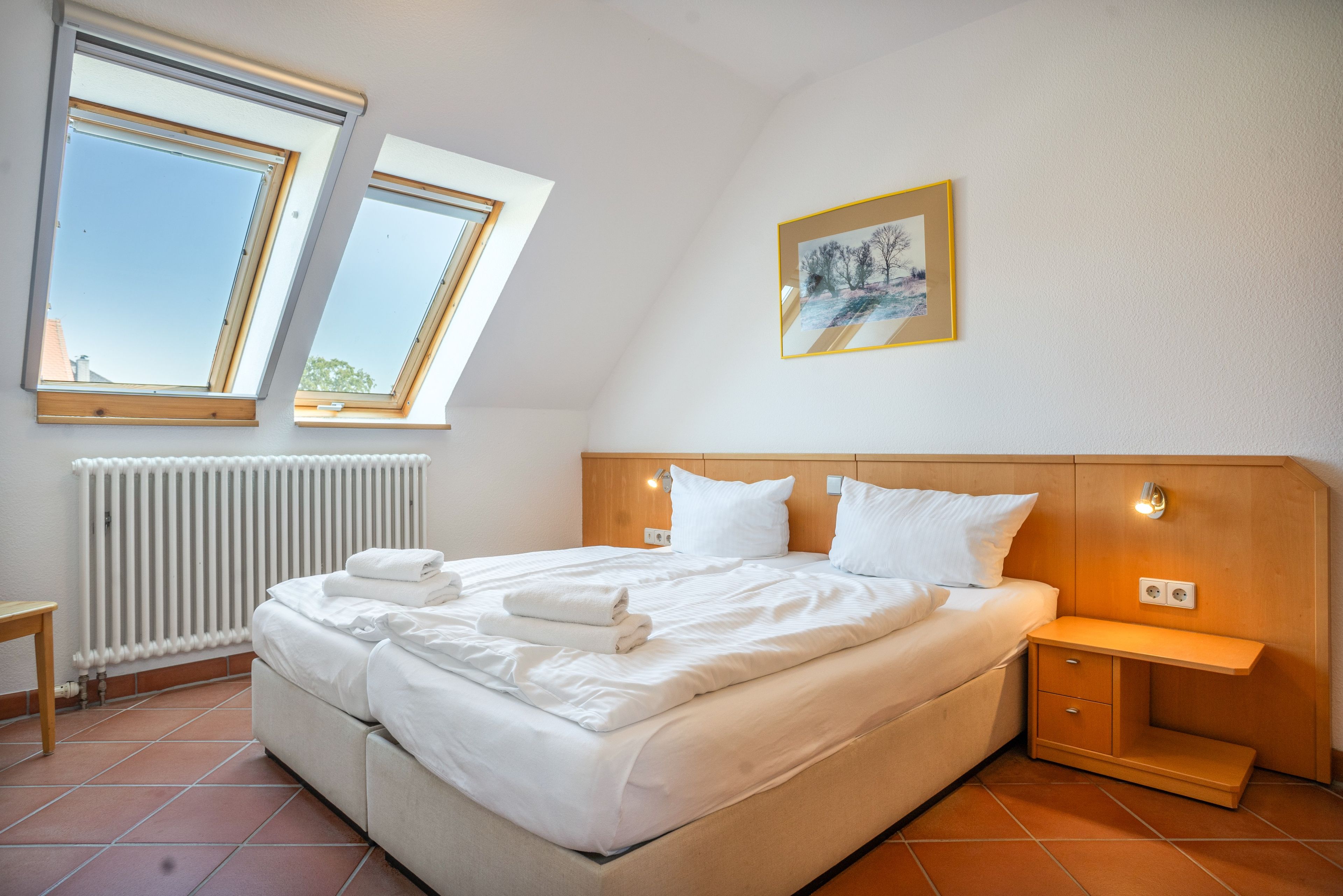 Una habitación de hotel con una cama grande, una mesita de noche, un radiador y dos ventanas.