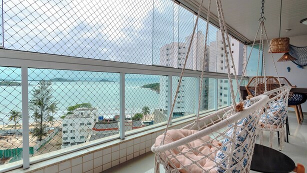 A room with a large window overlooking a beach, a hanging chair, and a dining area with chairs. - Blue Mar Apartamento (Guarapari)