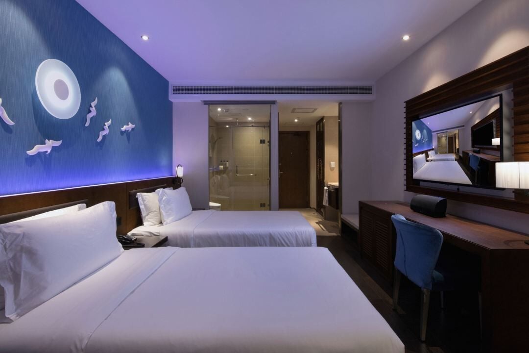 Habitación de hotel con dos camas, un escritorio, un televisor y un baño visible a través de una puerta de vidrio.