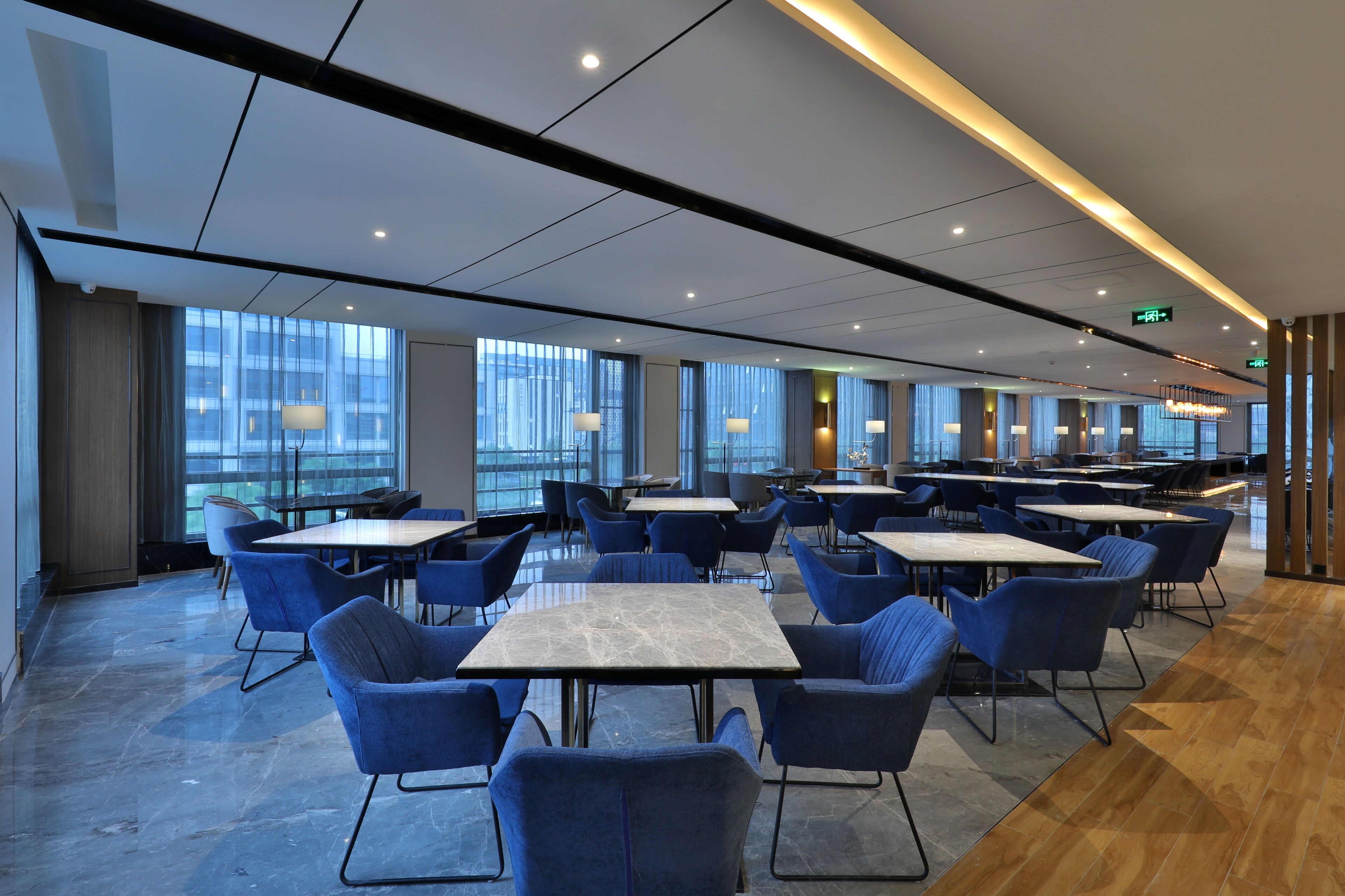 A spacious indoor area with tables and chairs arranged for dining, large windows allowing natural light, and a modern ceiling design.