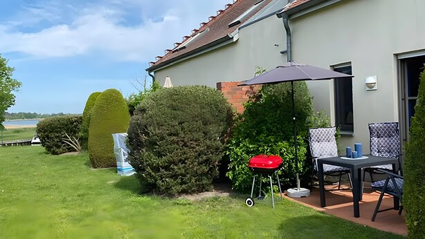 Eine Terrasse mit Tisch und Stühlen, ein roter Grill und Blick auf einen See.