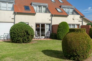 Een tweelaags huis met een rood pannendak, een overdekte patio met een tafel en stoelen, en verzorgd gesnoeide heggen.