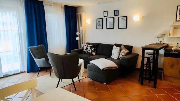 Ein modernes Wohnzimmer mit einer Couch, Sesseln, einem kleinen Tisch und einer Küchenzeile mit einer Arbeitsplatte und Barhocker.