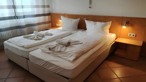 Una cama doble con ropa de cama y toallas blancas, cabecera de madera, lámparas de noche y una mesita de noche con un cajón.
