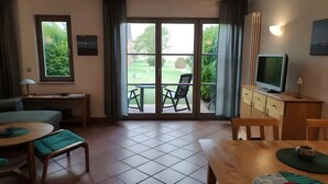 Una habitación con una mesa de comedor, sillas, televisor, sofá y vistas a un jardín a través de un ventanal.