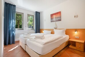 A hotel room with two beds, a view of trees through the windows, a painting of a sunset, and a small table with a lamp. - “Krienker See” Apartment – Terrace & Nature Views (Mellenthin)