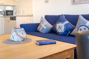 Une table en bois ornée d’un vase décoratif et d’un livre, un canapé bleu avec des coussins à motifs, et une cuisine en arrière-plan.