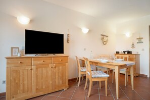 A dining area with a wooden table and chairs, a television mounted on the wall, and a kitchen area with a stove and microwave. - “Krienker See” Apartment – Terrace & Nature Views (Mellenthin)