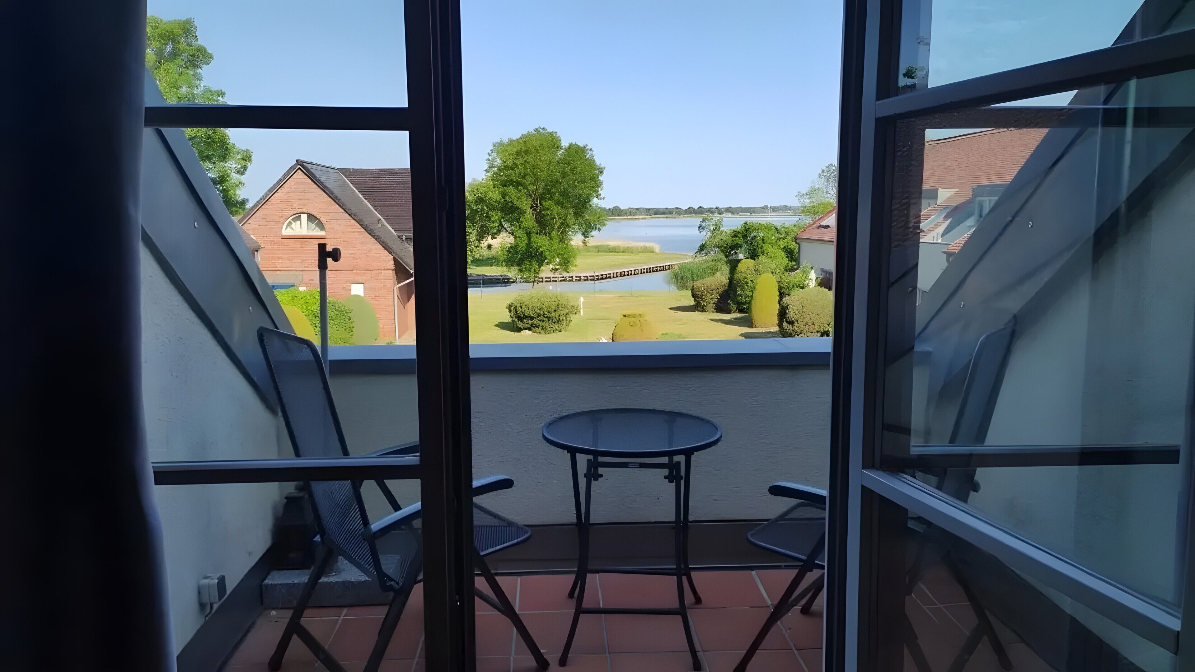 Un balcon avec une table et des chaises, donnant sur une étendue d’eau et sur de la verdure.