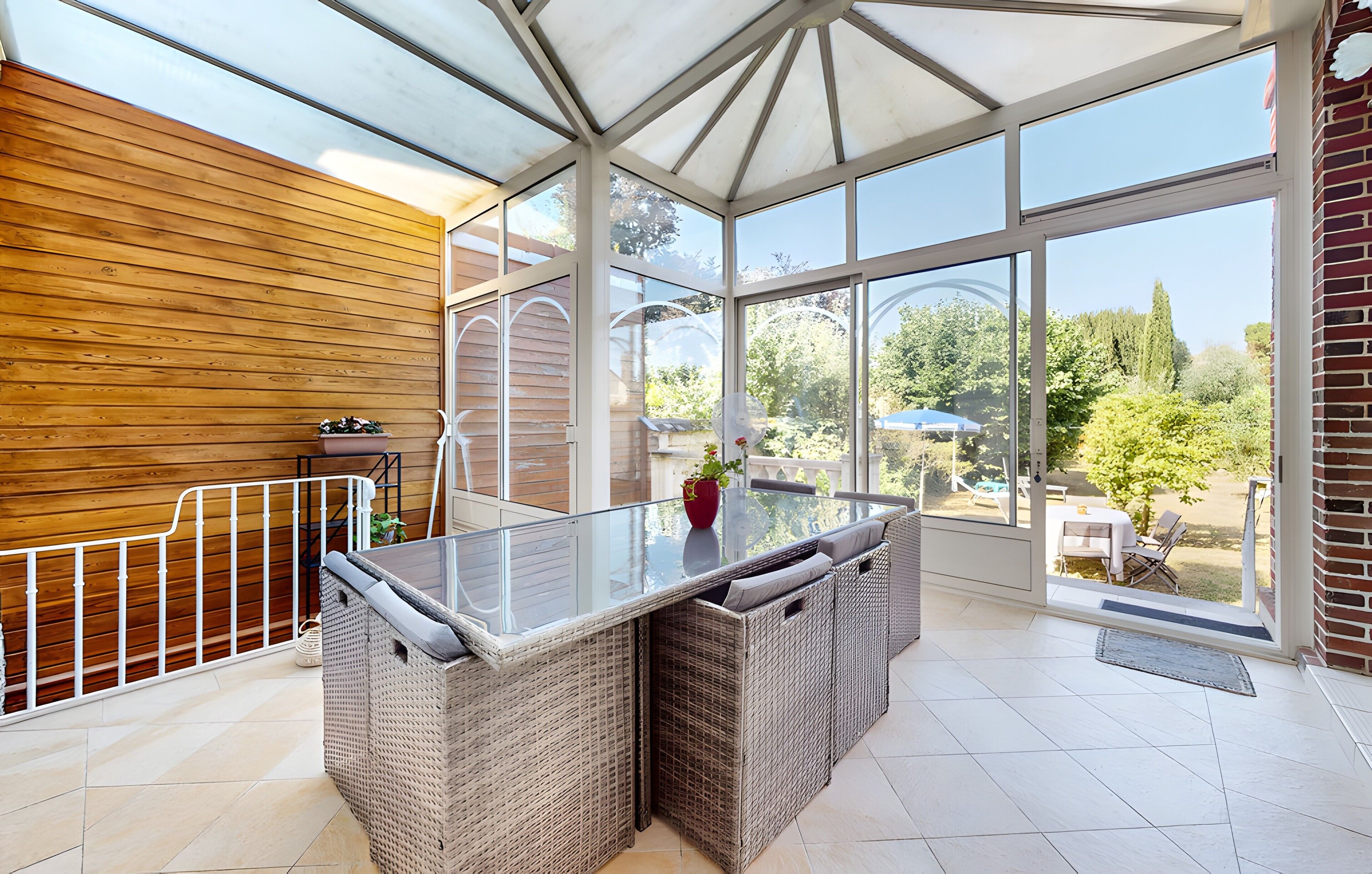 A covered patio area with a glass table and wicker furniture, a wooden wall, and a view of the garden.
