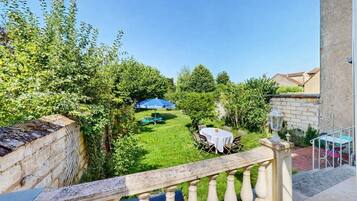 A balcony with a view of a well-maintained garden, a table with chairs, and a blue umbrella.