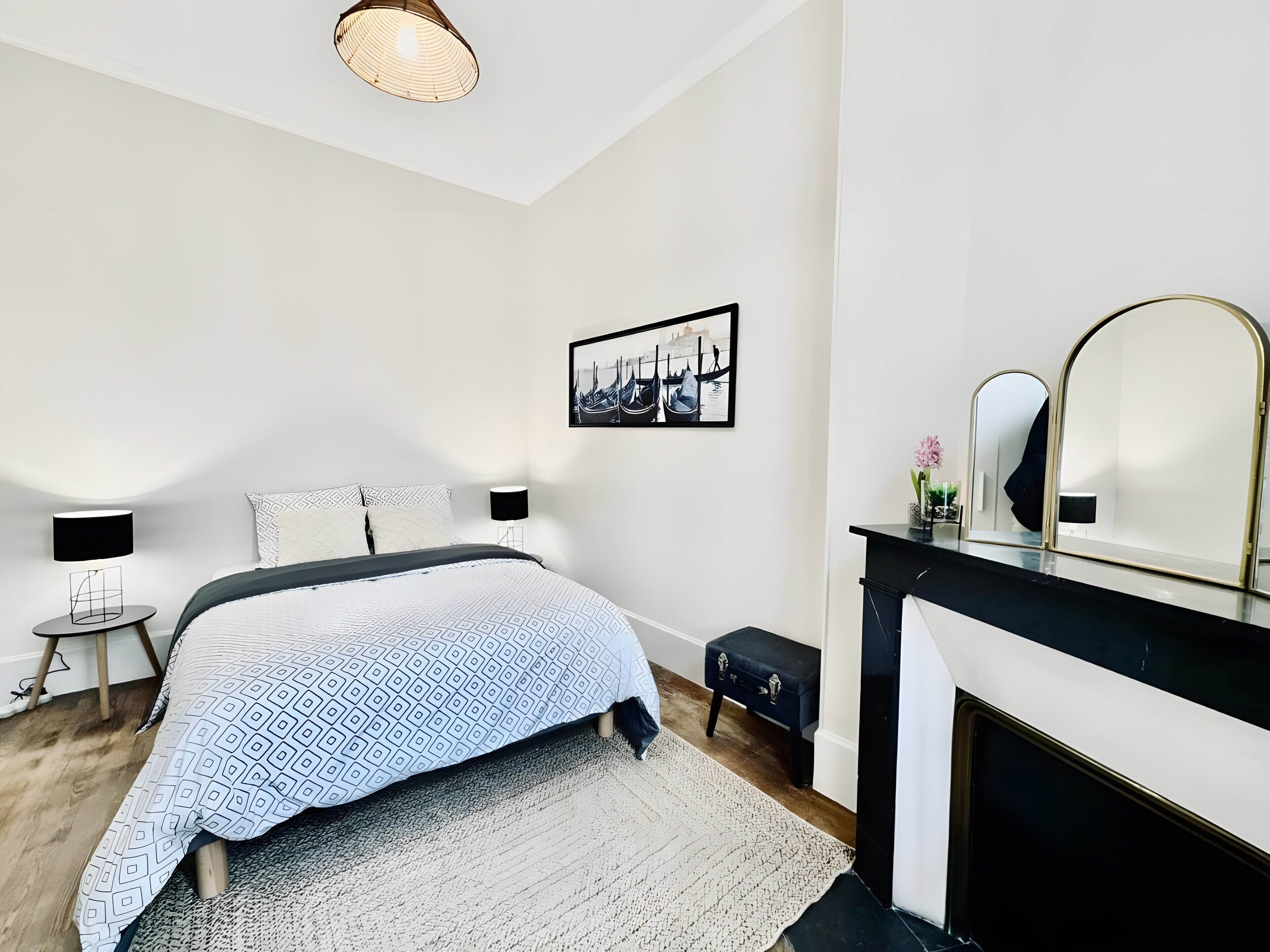 A bedroom with a bed, bedside lamps, a fireplace, a mirror, and a wall-mounted picture.