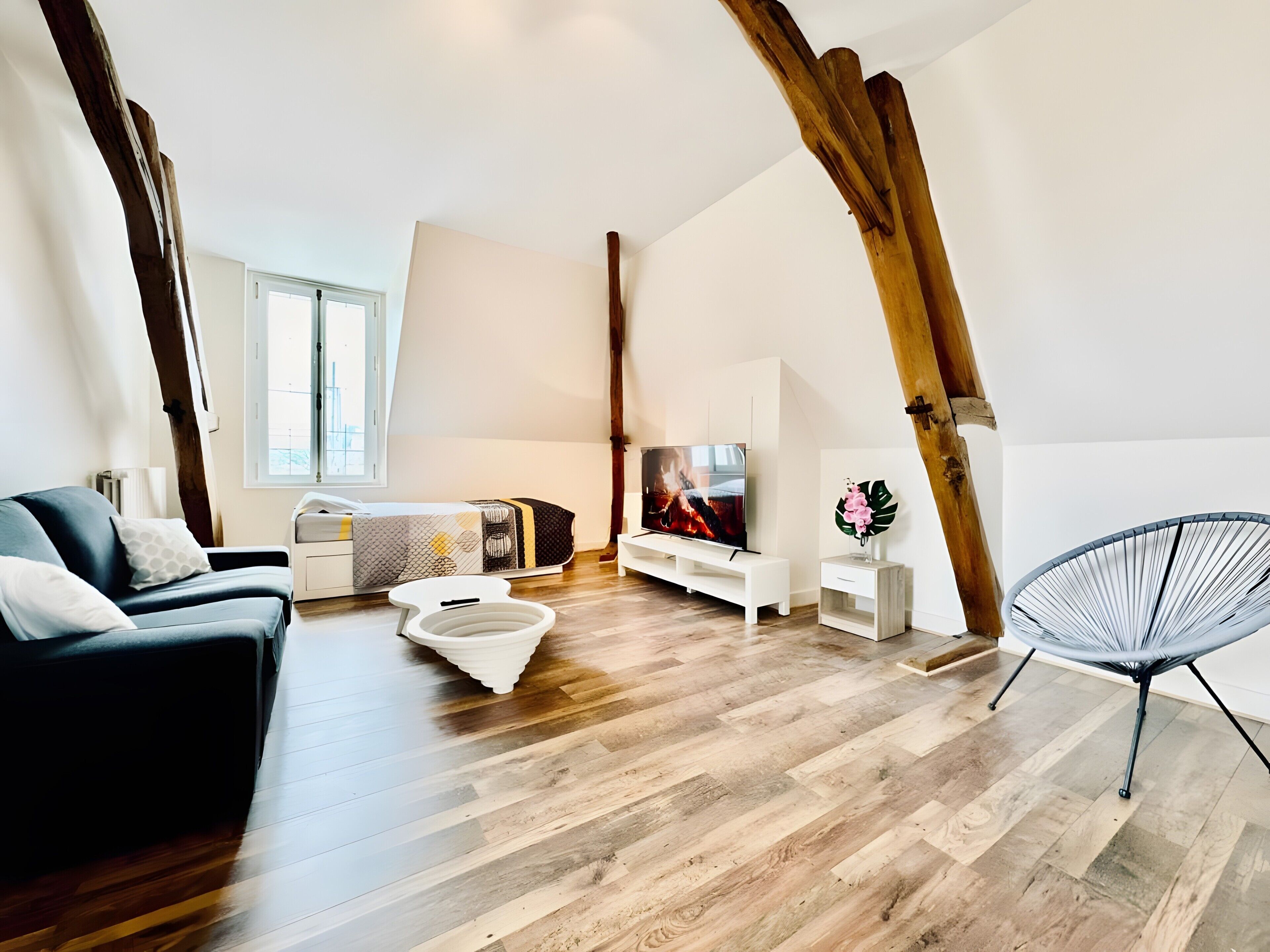 A modern loft-style room with exposed wooden beams, a black sofa, a white bench, a TV, and a hanging chair.