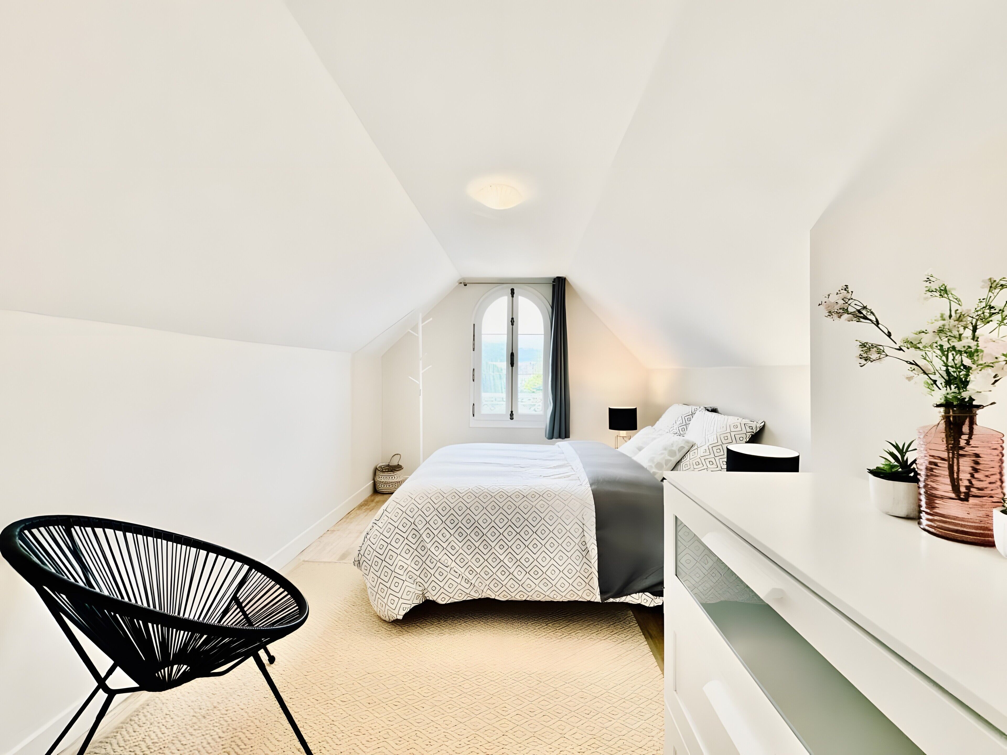 Une chambre à coucher avec un lit, une chaise, une table de chevet, une lampe et une fenêtre.
