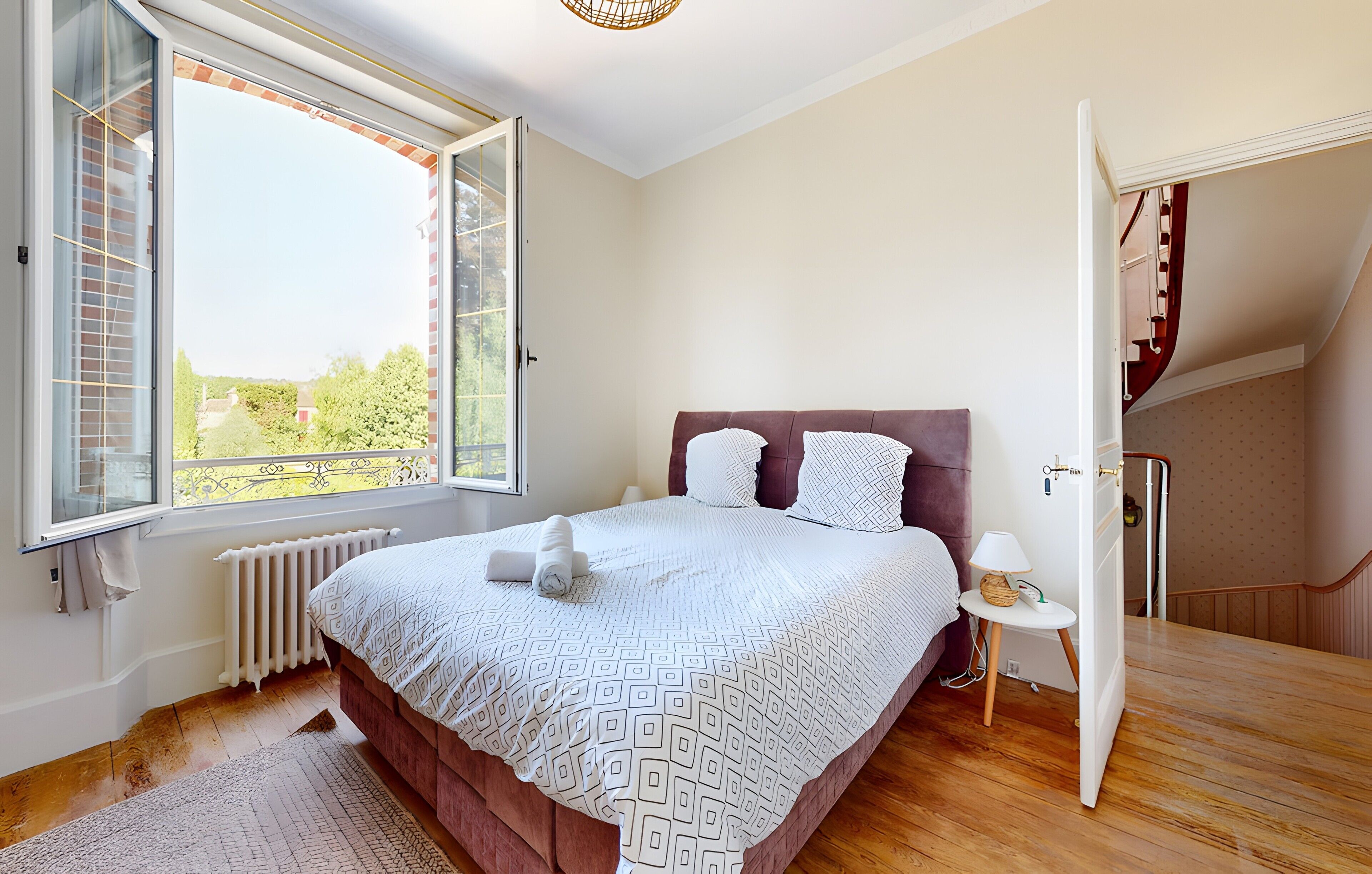 A bedroom with a bed, a window, a radiator, a bedside table with a lamp, and a staircase.