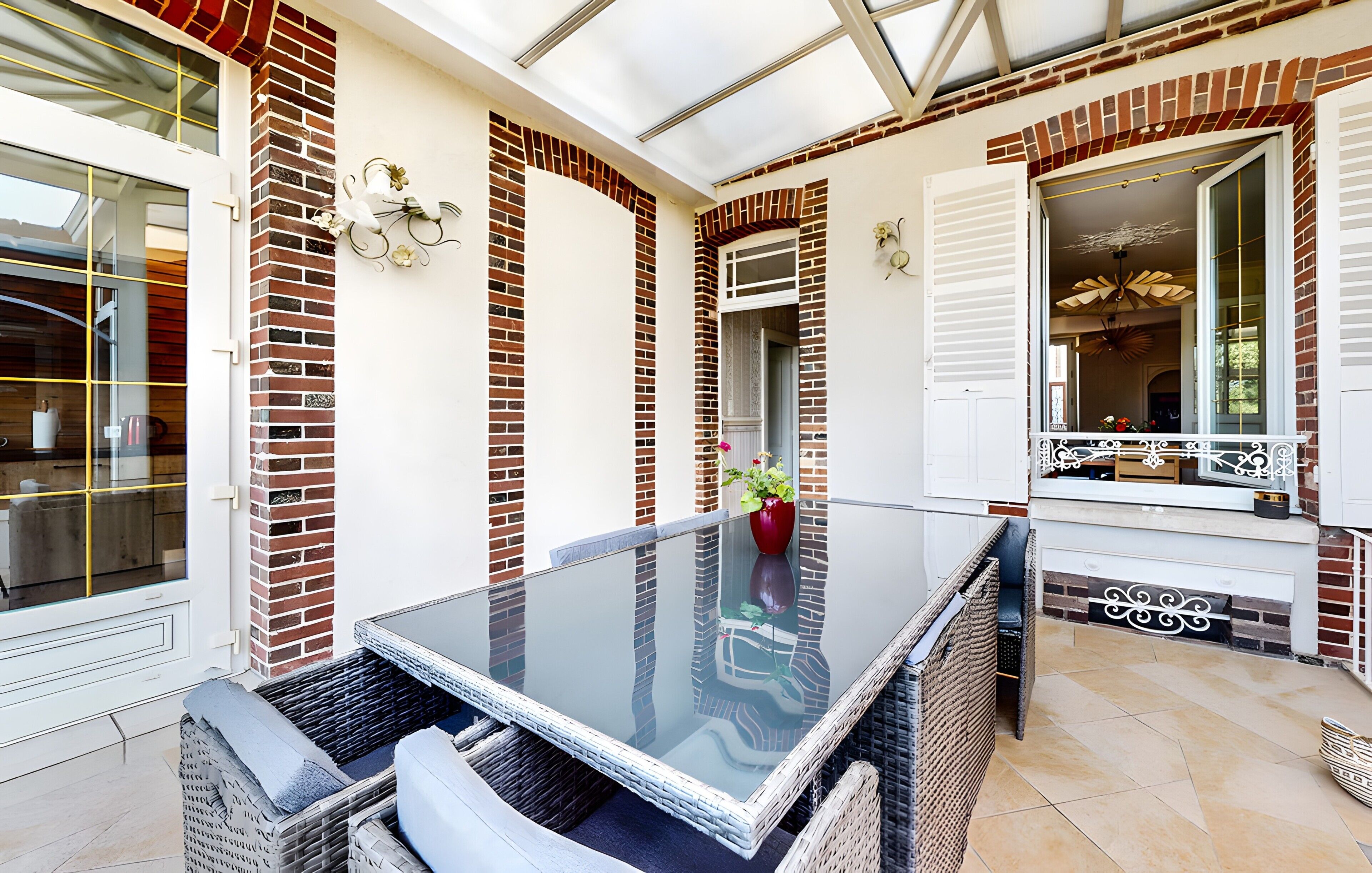 A patio with a glass table, wicker chairs, and a red vase with flowers.