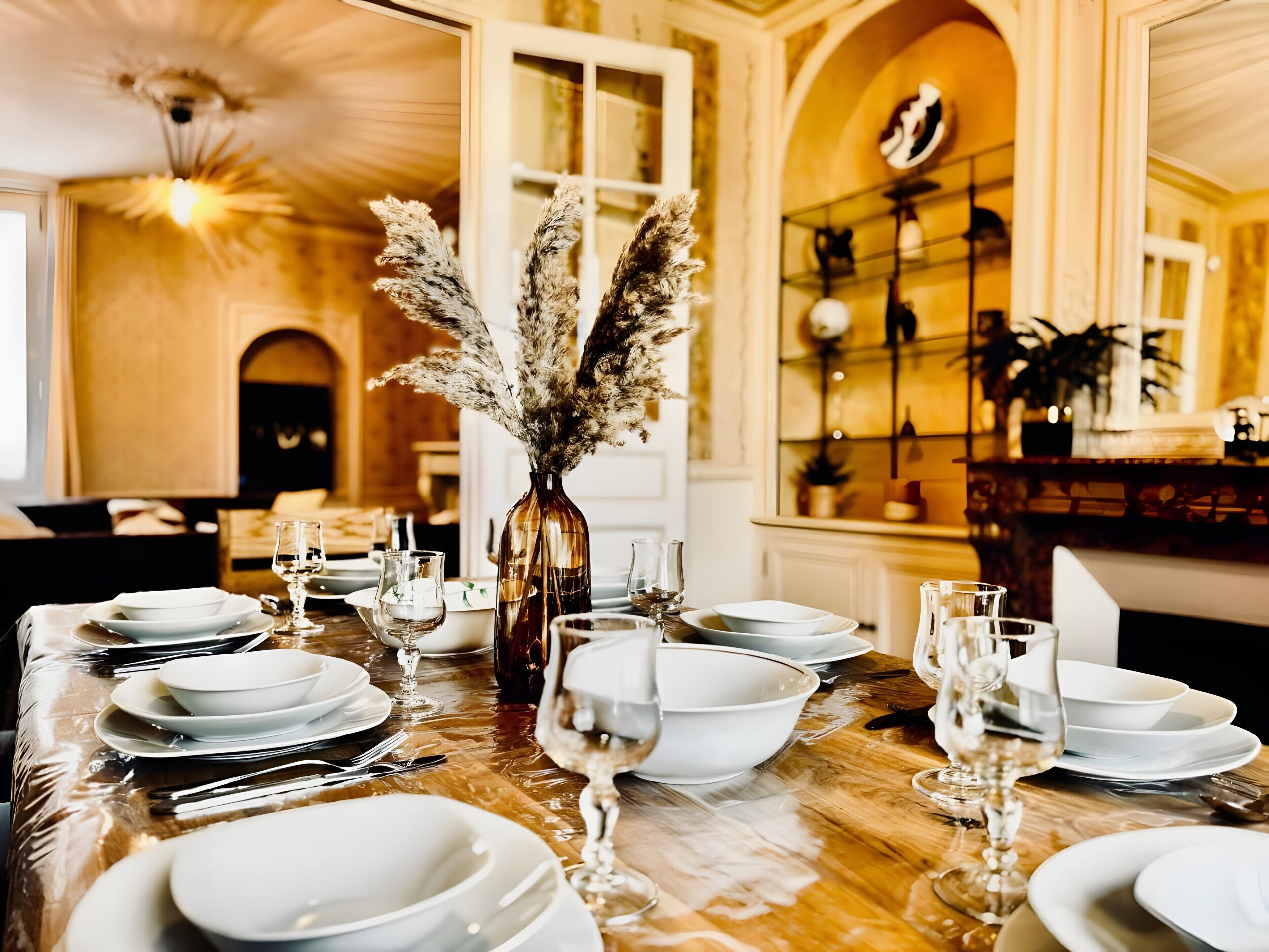 A dining table set with white plates, glasses, and cutlery, a vase with dried pampas grass, and a chandelier.