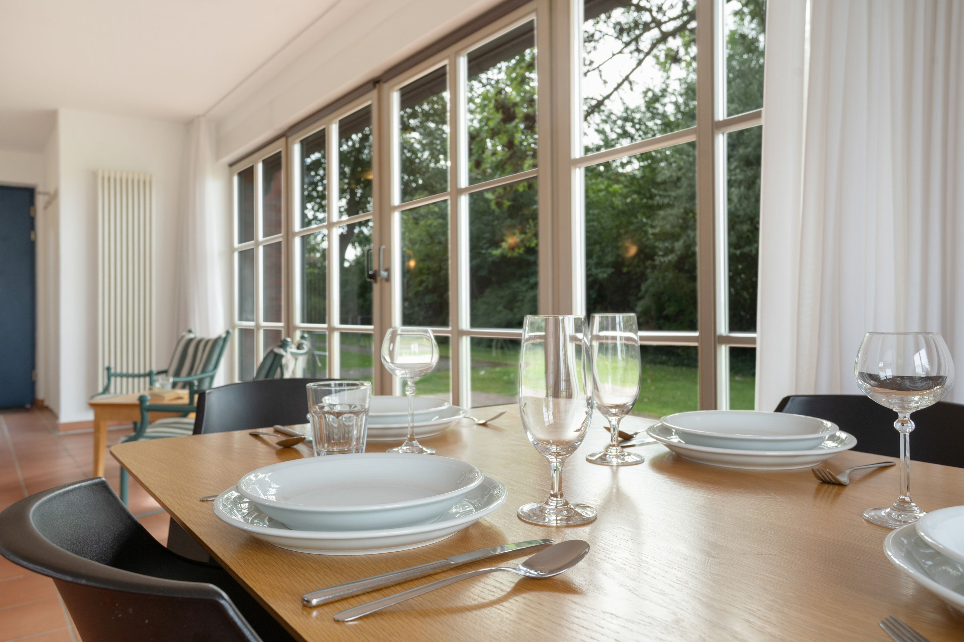 A dining table set with plates, glasses, and cutlery, with a view of greenery through large windows.
