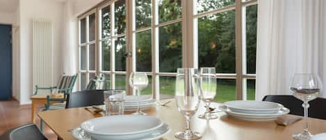 A dining table set with plates, glasses, and cutlery, with a view of greenery through large windows.