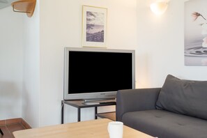 Ett vardagsrum med en platt-TV, en grå soffa, ett träbord och en inramad tavla på väggen.