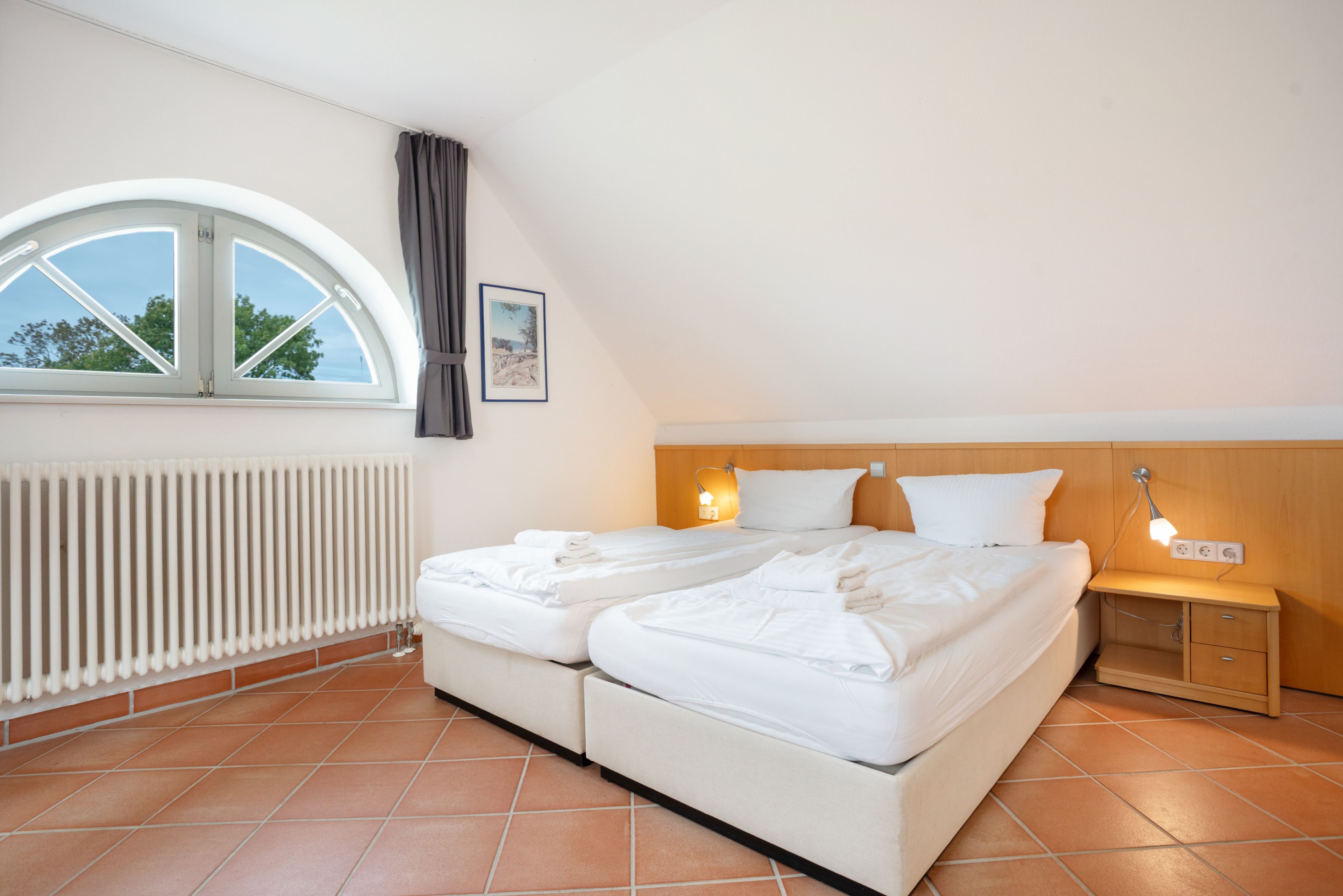 A bedroom with a double bed, a bedside table, a nightstand, a window with a view, and a radiator.