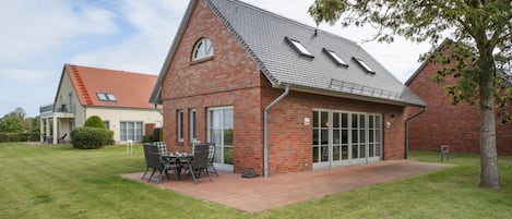 Una casa de ladrillo de dos pisos con techo inclinado, un patio con mobiliario exterior y un ventanal grande.