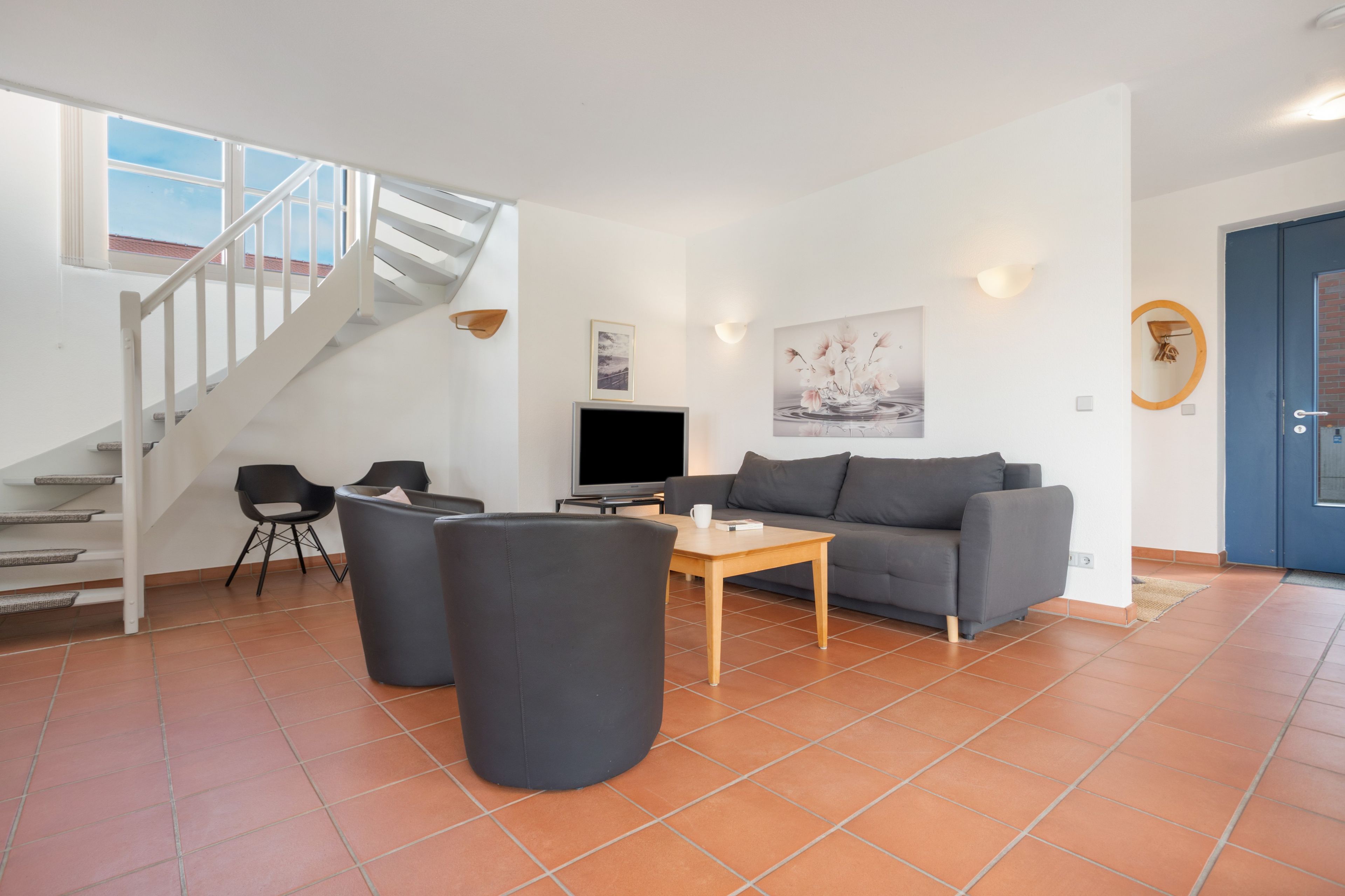 Ein modernes Wohnzimmer mit einem grauen Sofa, einem Couchtisch und einem Fernseher. Eine Treppe mit weißen Geländern führt zu einer oberen Etage.