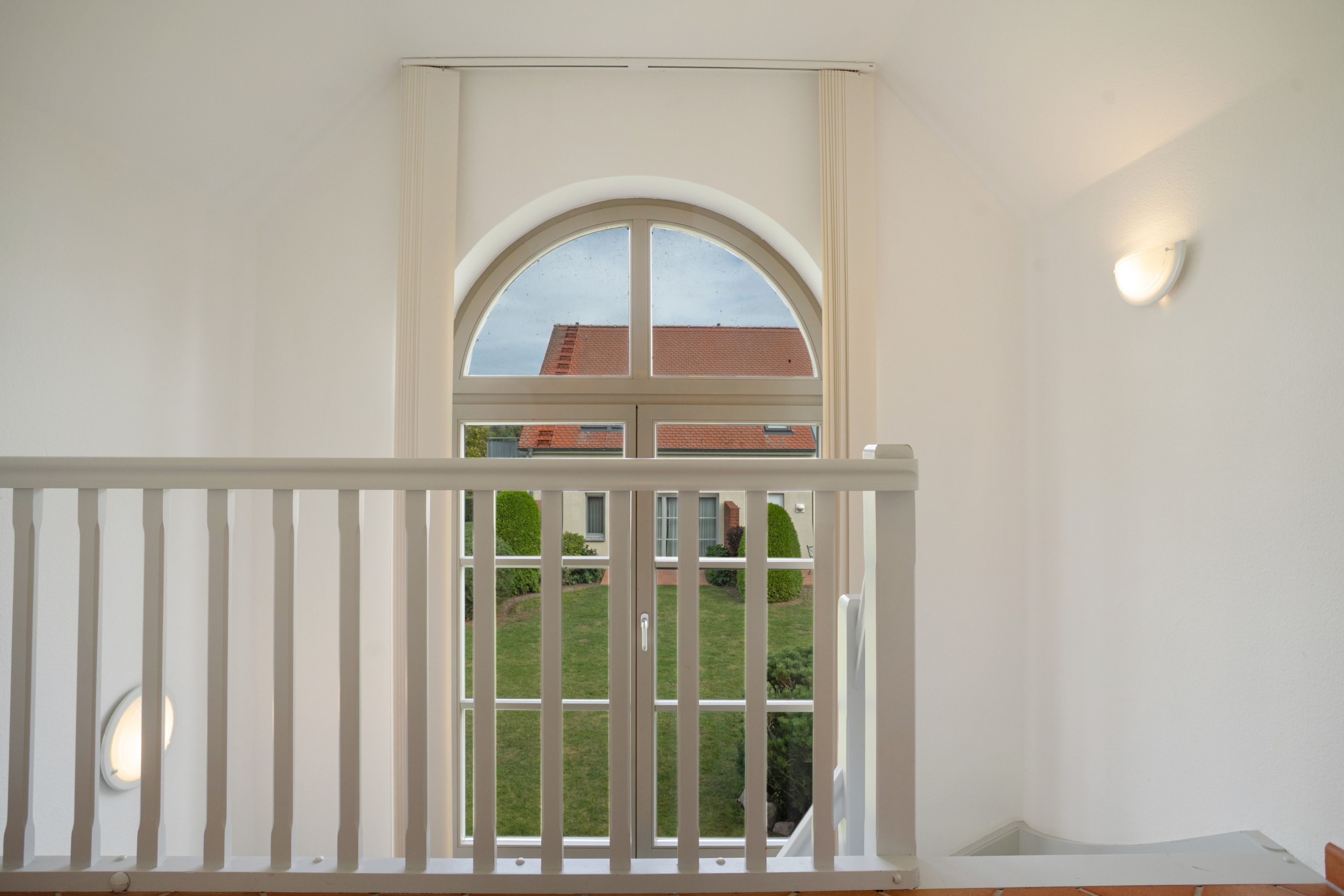 Ein Zimmer mit weißer Geländer, einem großen Bogenfenster und Blick auf ein Gebäude mit rotem Dach.