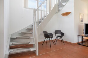 Un escalier avec des rampes blanches et une vue sur le ciel par les fenêtres. On y trouve deux chaises noires et un téléviseur posé sur un meuble.