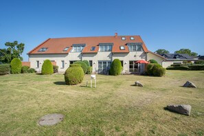 A two-story house with a red-tiled roof, multiple windows, and a well-maintained lawn with a manhole cover, rocks, and trimmed bushes. - "Kranich" – Bright Holiday Apartment for Nature Lovers in Usedom's Achterland (Mellenthin)