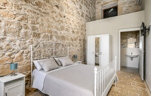 Una camera da letto con parete in pietra, un letto con struttura in metallo bianco, un armadio e un bagno con lavandino e specchio.