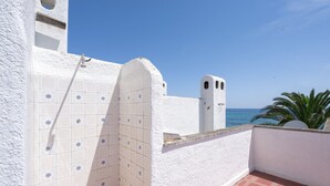 Un balcon carrelé de blanc, avec un sol recouvert de carreaux rouges, un mur blanc percé d’une fenêtre et une vue sur la mer.