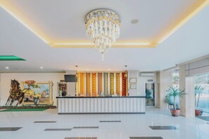 Vestíbulo de hotel con mostrador de recepción, una araña y un mural de un carruaje tirado por caballos, además de plantas en macetas.