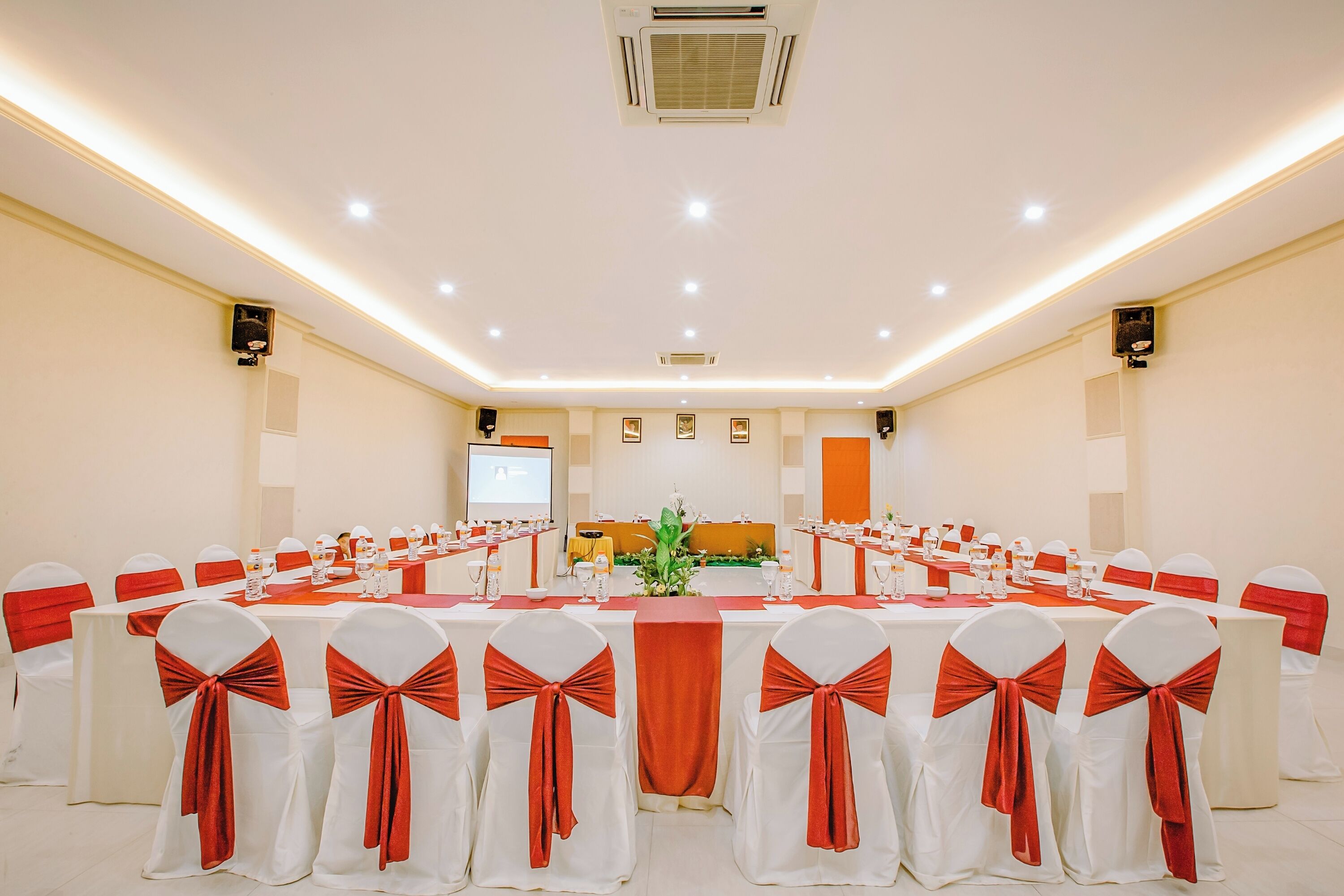 Un salón de conferencias con una larga mesa, sillas blancas adornadas con cintas rojas, una pantalla de proyector y altavoces.